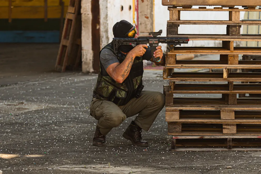 Jogador de balaclava agachado atrás de uma pilha de paletes, arma apoiada no ombro, mirando o pátio.