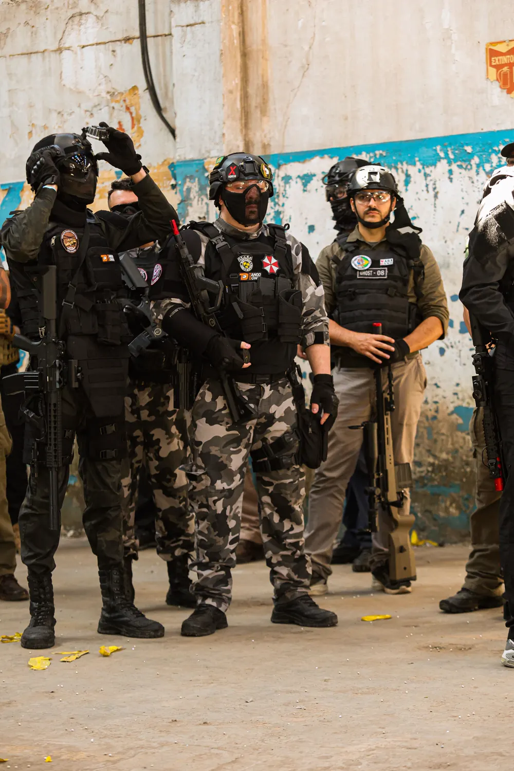 Grupo de jogadores reunido dentro do galpão durante o briefing, com equipamentos e camuflagens variados.