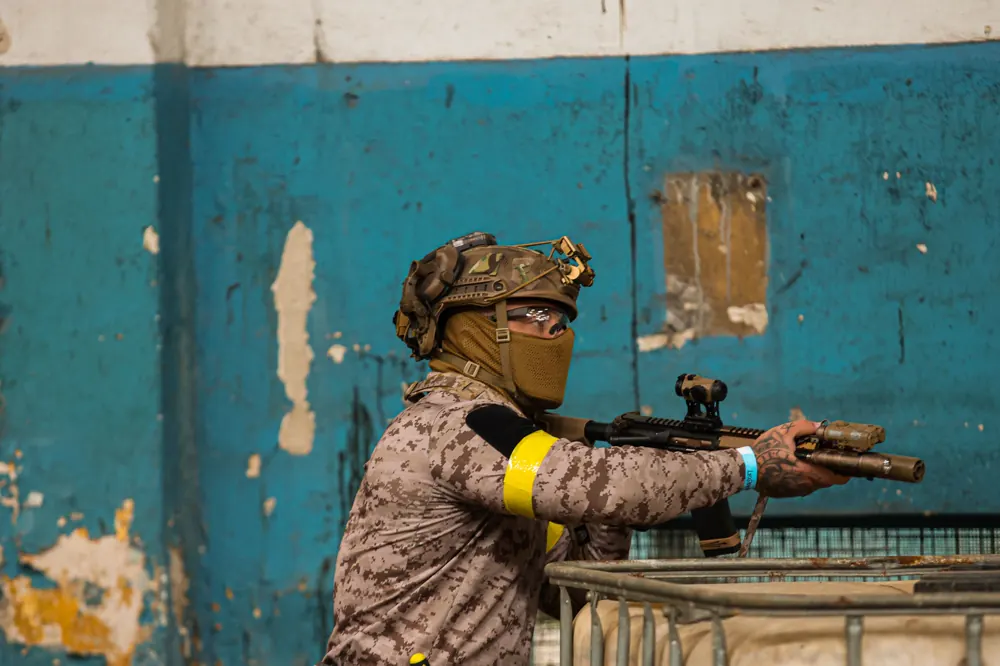 Operador de camuflagem deserto apoia a arma numa grade, recortado contra uma parede azul descascada.