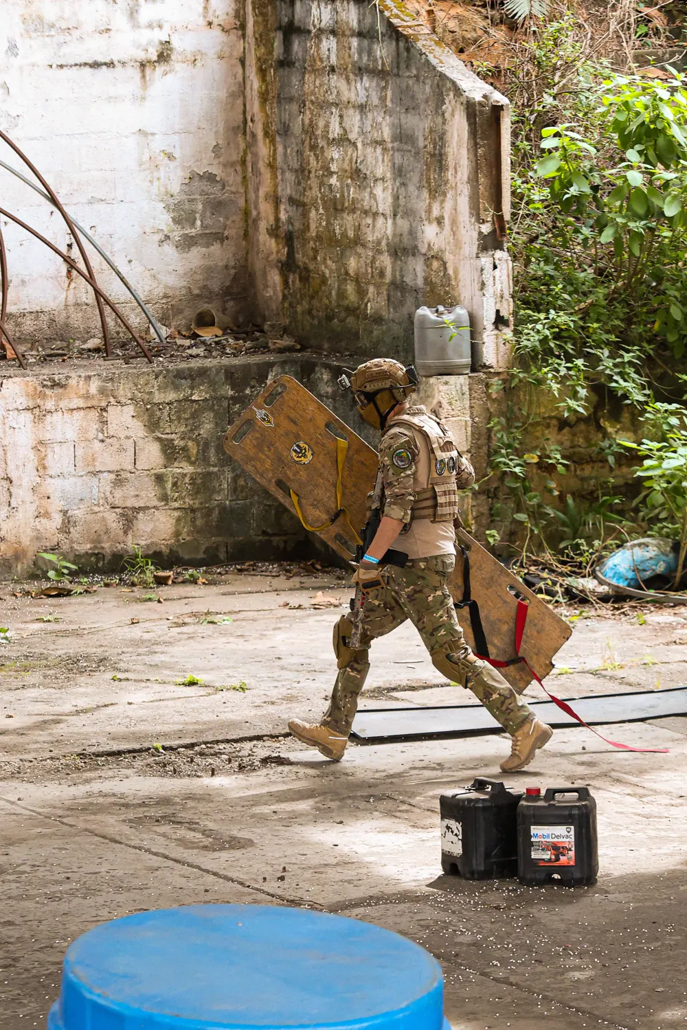 Operador em camuflagem atravessa o pátio carregando um escudo de madeira, entre paredes tomadas por mato e galões espalhados pelo chão.