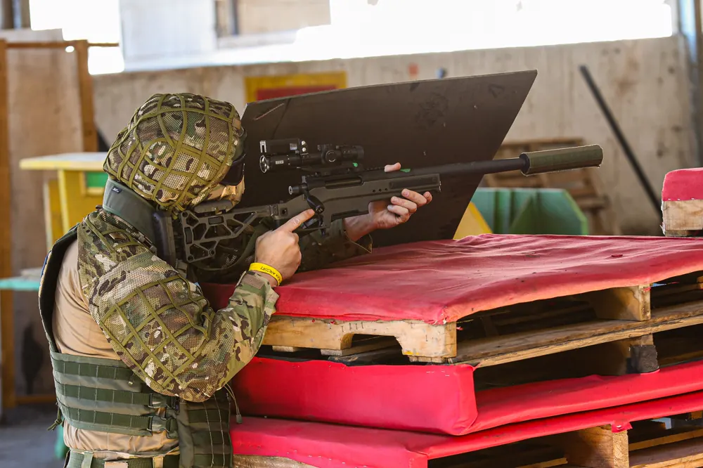 Sniper camuflado apoia o rifle sobre paletes cobertos por colchonetes, na área coberta do campo.