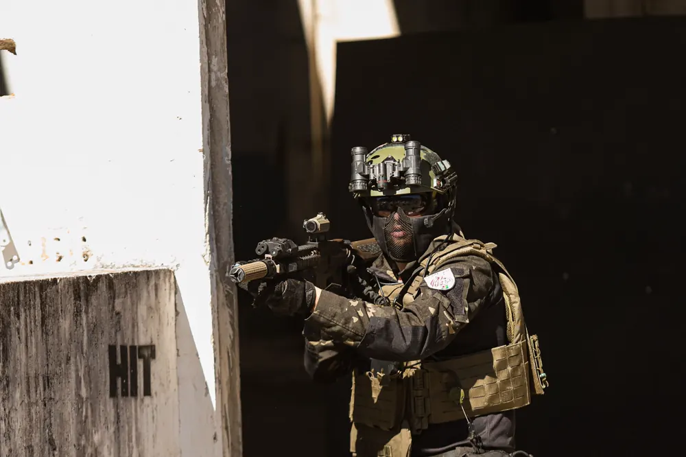 Jogador equipado avança ao lado de um muro com a palavra HIT pintada, arma na mira.