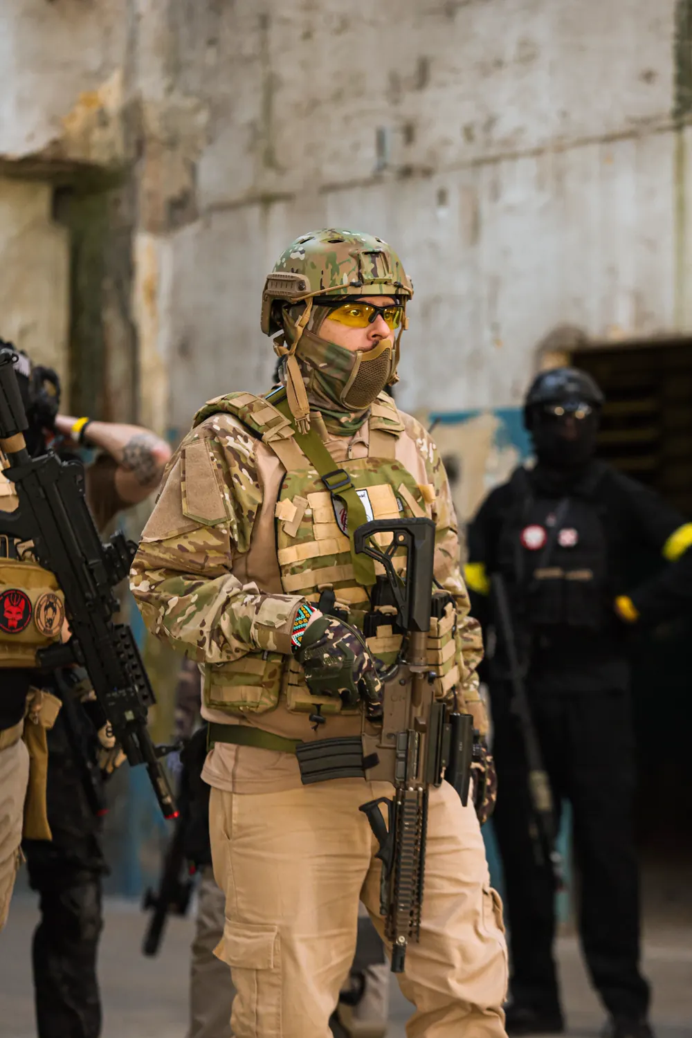 Operador de camuflagem multicam e óculos laranja em pé dentro do galpão, com dois jogadores de preto ao fundo.